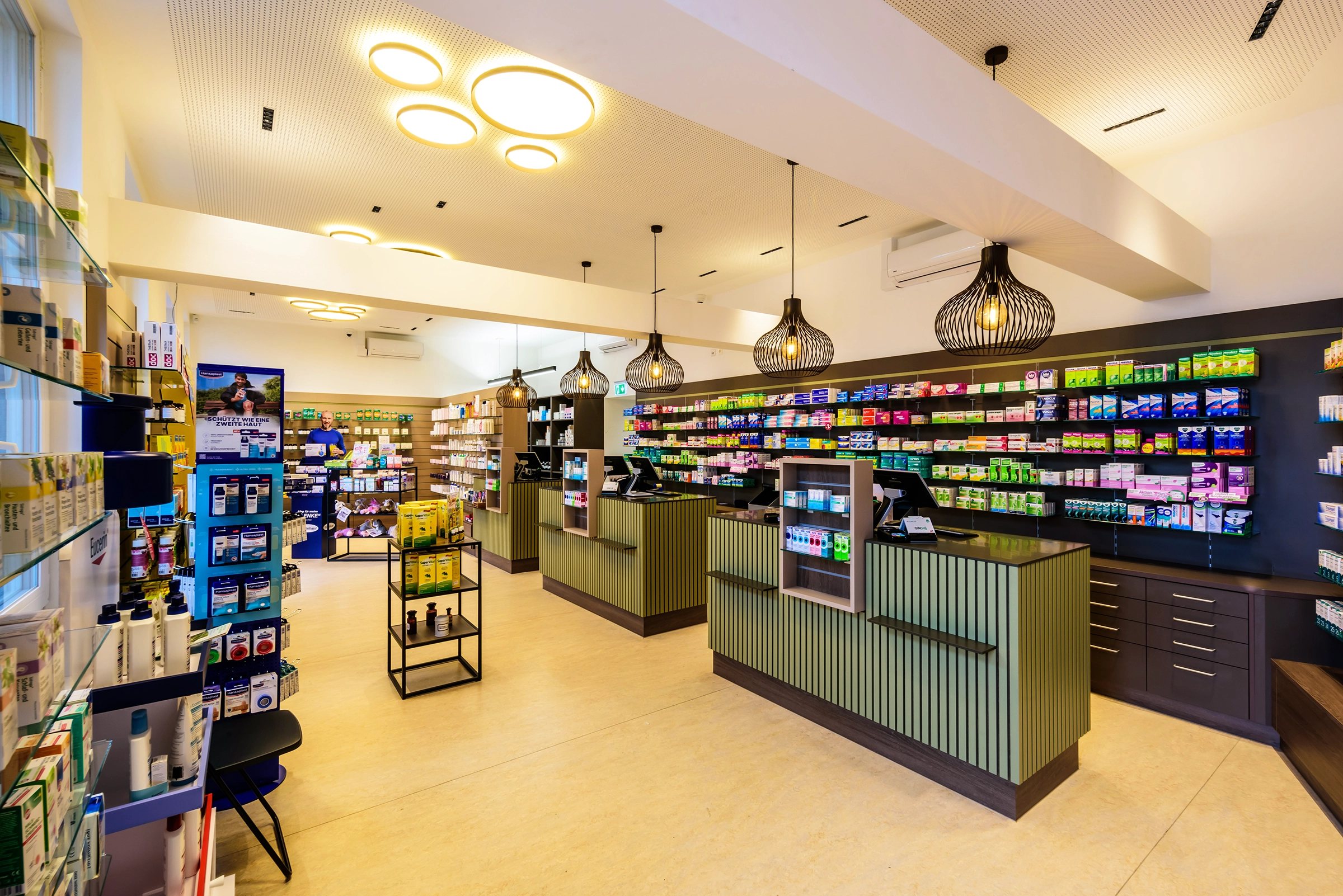 Blick in die Offizin der Mariahilf-Apotheke mit grünen Lamellentheken, Pendelleuchten und Regalwand