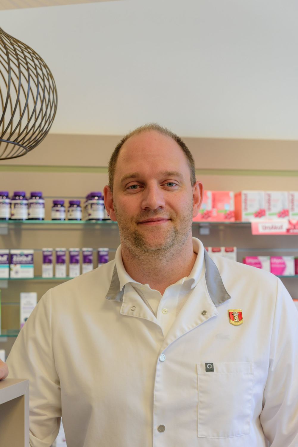 Mag. pharm. Florian Pfeiffer im weißen Kittel vor einem Regal der Mariahilf-Apotheke